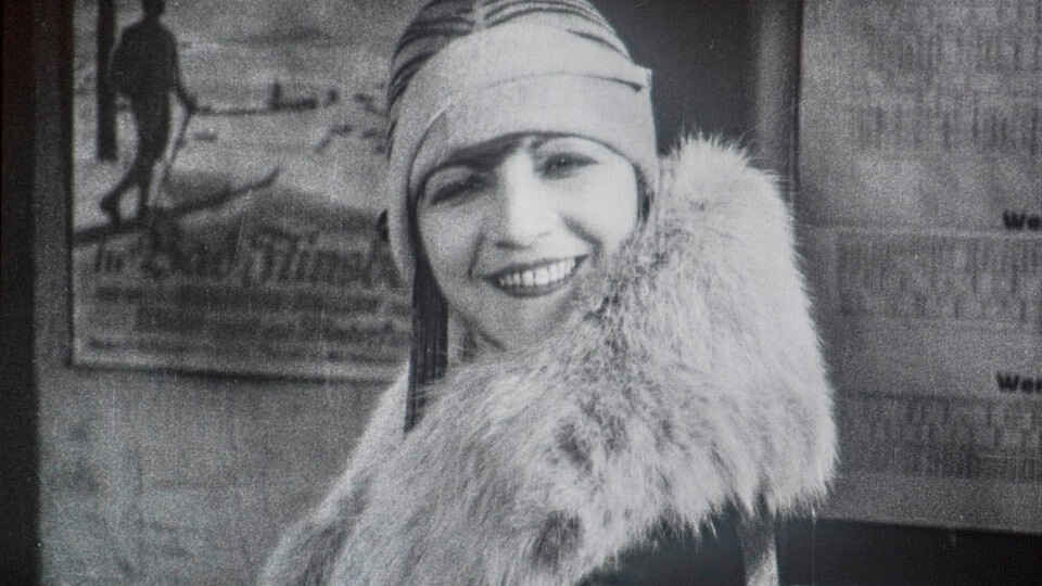 Ellen Richter smiling in a film close-up, wearing a striped cloche cap and a thick fur stole, with printed posters on the wall behind her