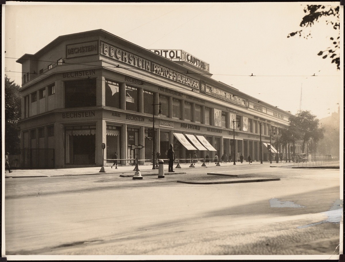 Photograph of a long low corner building with CAPITOL lettered on the roofline and Bechstein signage across the shopfronts below, seen across an almost empty street