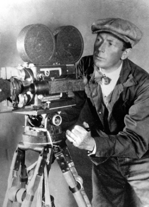 F. W. Murnau in a flat cap and work jacket, leaning in beside a hand-cranked film camera mounted on a wooden tripod