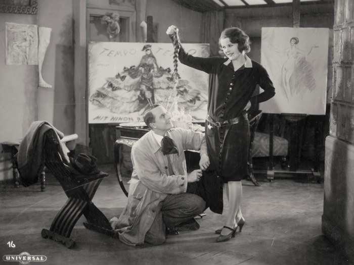 Publicity still from Moral: Ninon, in a dark belted coat and short bobbed hair, stands with one hand raised over a man kneeling at her feet in a scene-painter's studio, surrounded by costume sketches and a painted poster reading "Tempo Tempo" and "Mit Ninon d'Hauteville"